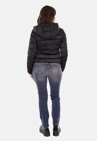 Giacca a vento nera con cappuccio e texture trapuntata, abbinata a jeans in denim blu e stivali neri grossi, vista da dietro.