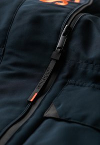 Giacca nera con accenti arancioni, caratterizzata da una zip prominente con la dicitura "SUPERDRY" e un tessuto strutturato, dal design elegante e moderno.