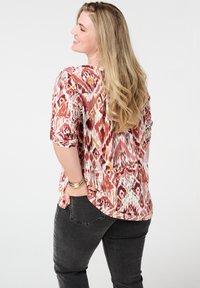 Multikleurige blouse met een rood-wit ikatpatroon, driekwartmouwen en een relaxte pasvorm, gecombineerd met donkergrijze jeans.