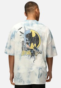 Camiseta de manga corta, tie-dye en blanco y azul claro, con un gráfico vintage de Batman que incluye murciélagos y un paisaje urbano. Material de algodón suave.