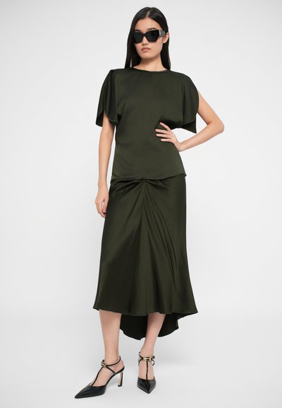 Abito verde oliva con maniche a farfalla corte, caratterizzato da un drappeggio frontale e un orlo asimmetrico. Abbinato a décolleté nere a punta.