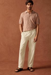 Brun tekstureret polo shirt, der er parret med lyse beige, plisserede, brede bukser og mørke sandaler. Enkelt design med en afslappet pasform.