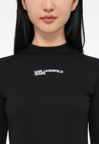 Detailní záběr na osobu v černém topu s vysokým límcem a na hrudi vyšitým logem "KARL LAGERFELD JEANS" v bílé barvě.
