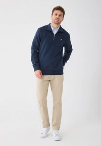 Tmavomodrá mikina na zips s golierom, s rebrovanými manžetami a lemom, v kombinácii s béžovými svetlými nohavicami a bielymi teniskami. Malý žltý detail loga.