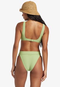 El conjunto de bikini de color verde claro con textura ribeteada cuenta con una parte superior de espalda baja con cierre ajustable y una parte inferior de cintura alta. Se lleva con un sombrero de paja tipo cubo.