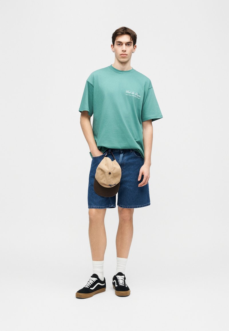 Joven de pie con camiseta oversize verde azulado, pantalones cortos vaqueros, calcetines blancos y zapatillas negras, sosteniendo una gorra beige y marrón a un lado.