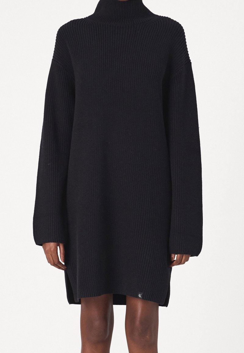 Robe pull en maille côtelée noire à col roulé, manches longues, fentes latérales et petit logo près de l'ourlet, portée par une personne avec mains et jambes visibles.