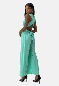 Pantalón corto mint con espalda descubierta, tela drapeada, piernas anchas y un detalle anudado en la cintura. Combinado con tacones altos negros.