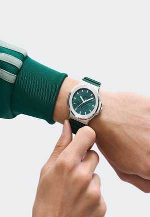 Person justerar en grön klocka med en silverram, iförd en grön långärmad topp med ljus- och mörkgrön rand på ärmslutet.