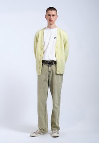 Ljust gult stickat kofta, vit t-shirt, gröna denimbyxor, grå sneakers. Modellen står mot en enfärgad bakgrund. Enkla designelement.