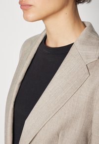 Beige bleiseri, jossa on rakenteellinen kaulus, teksturoitu kangas ja terävät hihat, mustan t-paidan päällä. Lähikuva kauluksesta ja olkapäästä.