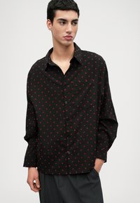 Camisa preta com bolinhas vermelhas, fecho de botões, mangas longas, um bolso no peito, tecido suave e corte descontraído.