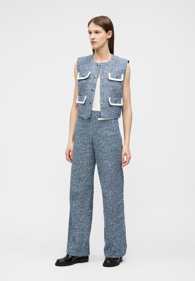 Γυναίκα με μπλε υφασμάτινο αμάνικο γιλέκο με λευκή λεπτομέρεια και ταιριαστό φαρδύ παντελόνι, συνδυασμένα με μαύρα παπούτσια, στέκεται μπροστά σε λευκό φόντο.