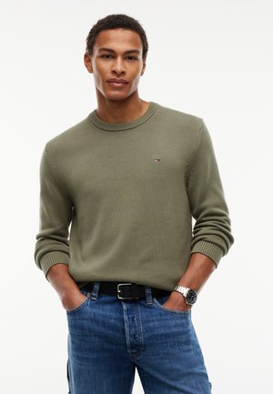 Mann trägt olivgrünen Pullover mit kleinem Logo, blaue Jeans, schwarzen Gürtel und silberne Armbanduhr, steht mit Händen in den Taschen.