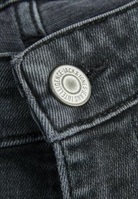 Primer plano de tela de mezclilla negra que presenta un botón de metal plateado con texto grabado y detalles de costura visibles a lo largo de los bordes.