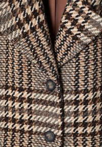 Houndstooth patterned coat in beige, brown, and black with a textured finish; features notched lapels and two dark buttons.