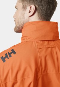 Oranžová voděodolná bunda s vysokým límcem a černým logem na rameni. Materiál má hladký, strukturovaný povrch.