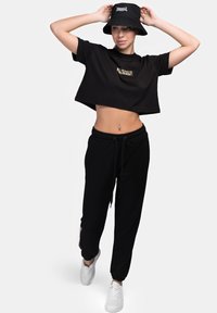 Black cropped t-shirt with a gold logo, black joggers with a drawstring, and a black bucket hat. White sneakers complete the outfit.