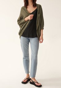 Olijfgroene open cardigan gelaagd over een zwarte top, gecombineerd met lichtblauwe spijkerbroek en zwarte teenslippers. Glad, lichtgewicht materiaal.