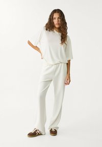 Bílé oversized tričko s krátkými rukávy, v kombinaci s volnými bílými kalhotami. Hnědé tenisky mají krémové akcenty. Hladká textura látky.