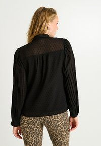 Blusa negra con tejido semitransparente de puntos, mangas plisadas y cuello abotonado. Combinada con unos vaqueros de estampado de leopardo.
