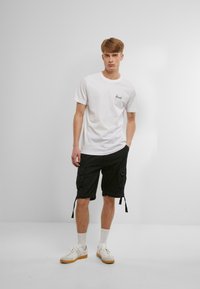 Hvid bomulds t-shirt med lille sort logo på brystet, parret med sorte cargo shorts med lommer og justerbare snore. Båret med hvide sneakers.