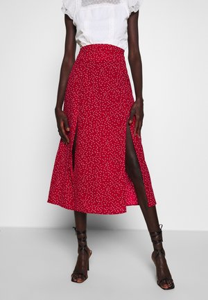 Jupe midi rouge à pois blancs, dotée d'une fente haute et d'une taille cintrée. La jupe est confectionnée dans un tissu léger, accompagnée de sandales à talons noires.