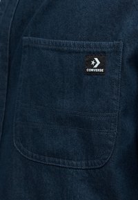 Σκούρο μπλε ύφασμα τζιν με τσέπη που φέρει μαύρο λογότυπο Converse με λευκό αστέρι και βέλος.