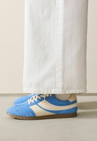 Blauwe suède sneakers met crèmekleurige leren accenten, voorzien van een gekarteld ontwerp, veters en een bruine rubberen zool, gecombineerd met witte spijkerbroeken.