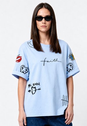 Femme portant des lunettes de soleil noires et un t-shirt bleu clair avec divers motifs graphiques noirs et rouges ainsi que des textes comprenant « faith » et « mi amor ».