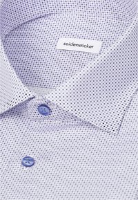 Gros plan d'un col de chemise à motifs avec des boutons bleus et une étiquette blanche indiquant le nom de la marque seidensticker.