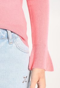 Vaaleanpunainen neulepaita, jossa on levenevät hihat, yhdistettynä vaaleansinisiin farkkuihin, joiden taskussa on kukkakirjontaa.