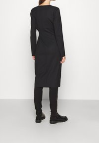 Schwarzes langärmeliges Kleid mit gerader Silhouette, hohem Kragen und einem Reißverschluss hinten. Kombiniert mit schwarzen taillierten Hosen und Stiefeletten.