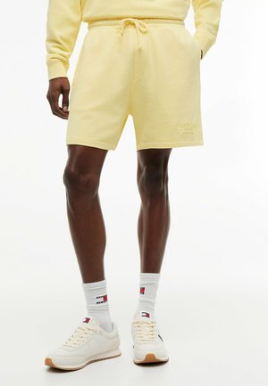 Man draagt lichtgele kortebroek met trekkoord, bijpassende trui, witte merk-sokken en off-white sneakers en staat.