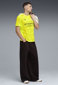 Gul sportströja med en strukturerad mönstring, med "ETIHAD AIRWAYS"-logotyp. Kombinerad med vida svarta byxor och vita slip-on skor.