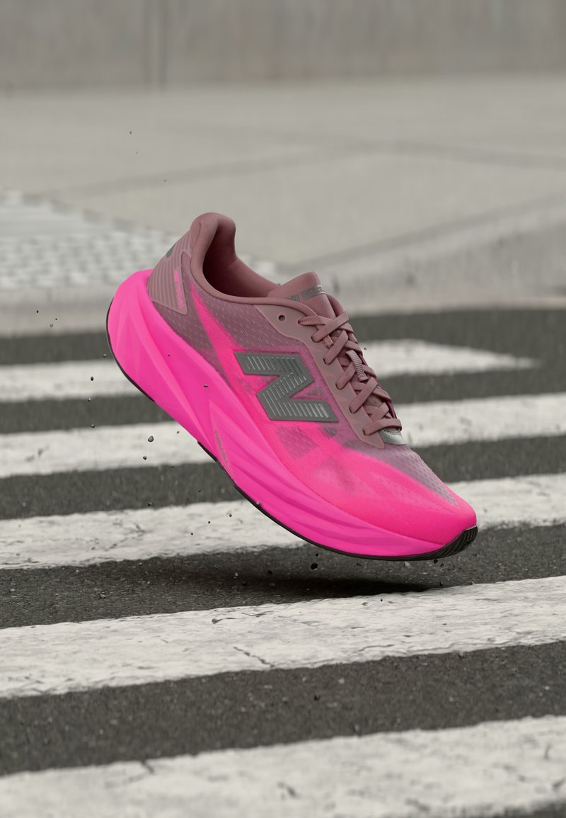 Chaussure de course rose vif et mauve avec un logo "N" gris, flottant au-dessus d'un passage piéton rayé noir et blanc sur une route en asphalte.