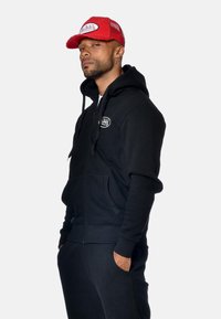 Uomo con barba e orecchini che indossa un felpa nera con zip e un cappellino rosso Van Dutch, in piedi con le mani nelle tasche su uno sfondo semplice.