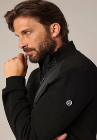 Zwarte zip-up jas met een gewatteerd paneel, geribde boorden en een logo-accent op de mouw. Glad materiaal met een aansluitende pasvorm.