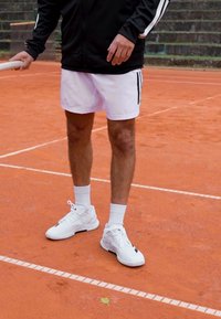 Valged tennisekad koos mustade aktsentidega, valged spordilühikesed püksid ja must tõmblukuga jakk valgete triipudega, clay tennisekohas.