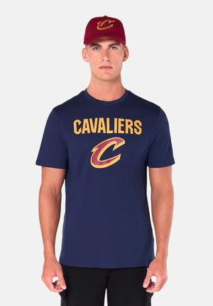 Man die een donkerblauw Cavaliers t-shirt draagt met een groot logo en een bijpassende bordeaux Cavaliers baseballpet, staand tegen een effen achtergrond.