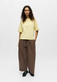 Femme aux cheveux bouclés portant un t-shirt ample jaune pâle, un pantalon large marron et des chaussures noires, debout avec une main dans la poche.