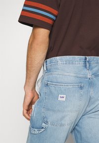 Vaqueros de mezclilla azul claro con corte holgado, con un bolsillo trasero y una pequeña etiqueta blanca. Camisa marrón de manga corta con rayas rojas y azules.