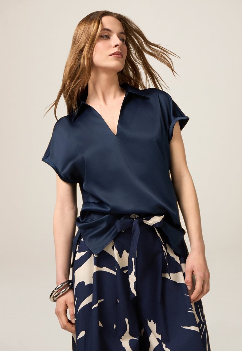 Bluză din satin bleumarin cu decolteu în V și mâneci scurte răsucite, purtată cu pantaloni largi cu un model abstract crem.