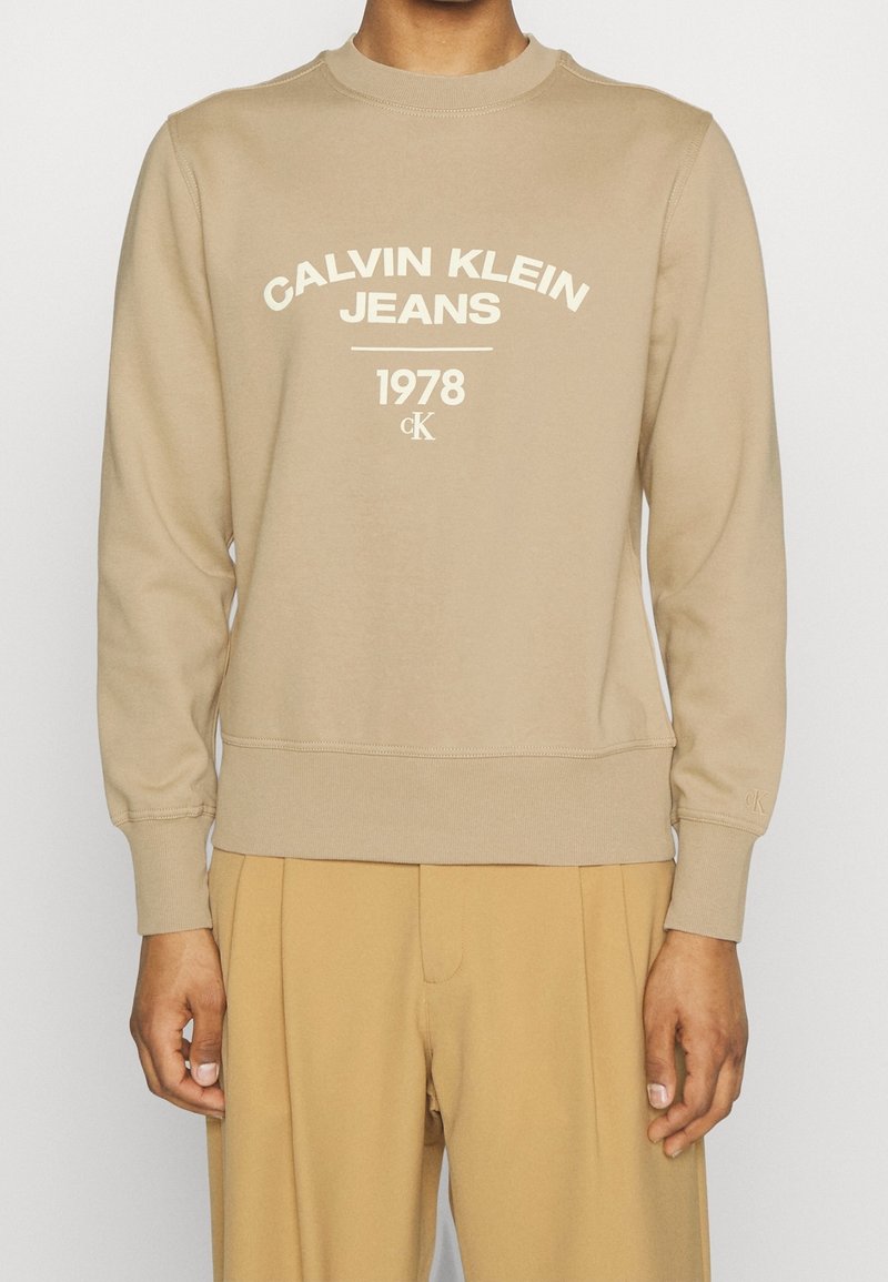 Beżowa bluza z długim rękawem z napisem "CALVIN KLEIN JEANS 1978 cK", noszona z dopasowanymi beżowymi spodniami, widok z przodu na środkową część ciała.