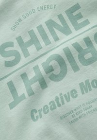 Jasnozielona tkanina z dużym tekstem "SHINE BRIGHT" i mniejszymi motywującymi frazami o dobrej energii i kreatywności.