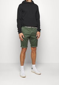 Felpa nera con cappuccio e tasca frontale, abbinata a pantaloni corti in cotone verde con orlo arrotolato e cintura a contrasto. Sneakers bianche.