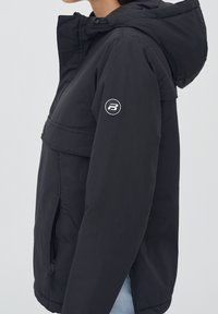 Chaqueta negra con capucha que presenta un cierre frontal con cremallera, bolsillos laterales y un logotipo circular en la manga izquierda. Fabricada con un tejido ligero y texturizado.