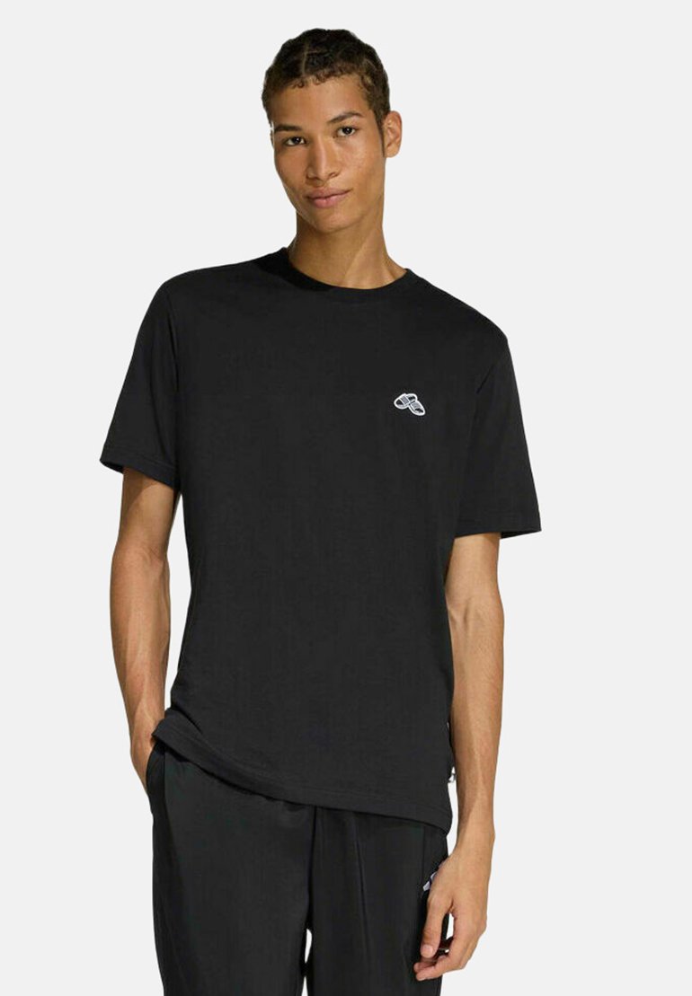 Jeune homme aux cheveux tressés portant un T-shirt noir avec un petit logo blanc sur le côté gauche de la poitrine et un pantalon noir, debout devant un fond uni.