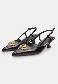 Črne lakirane slingback čevlje s koničastim nosom, majhnim peto in zlatim dekorativnim okovjem spredaj. Gladka tekstura.