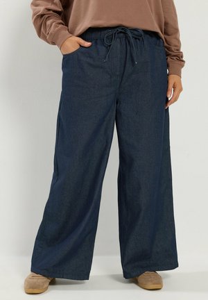 Personne portant un pantalon large bleu foncé avec taille à cordon et chaussures beiges, debout avec une main dans la poche et l'autre détendue.